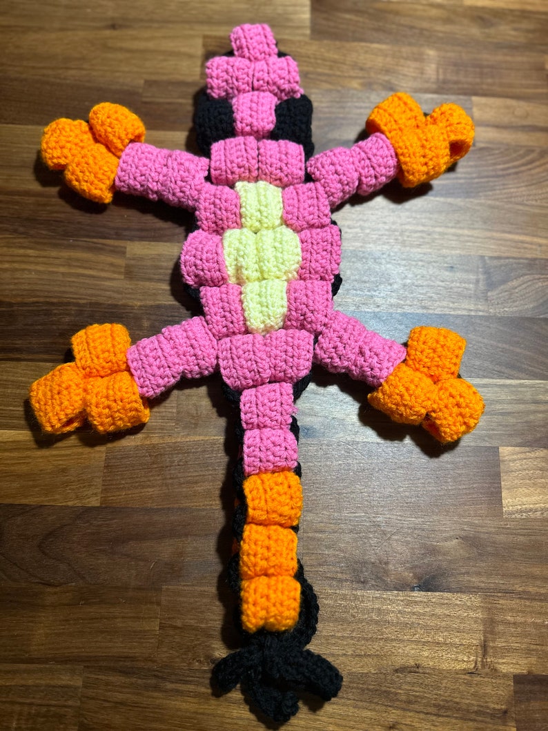 May include: A crocheted lizard toy with a pink, orange, yellow and black colour scheme. The lizard has a long tail and four legs.