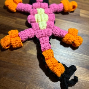 May include: A crocheted lizard toy made with pink, orange, yellow and black yarn. The lizard has a long tail and four legs.