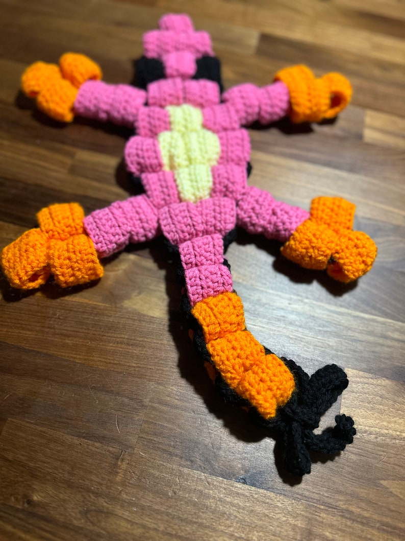 May include: A crocheted lizard toy made with pink, orange, yellow and black yarn. The lizard has a long tail and four legs.