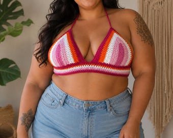 Pode incluir: Um top halter de crochê com listras horizontais em tons de magenta, laranja e branco, combinado com shorts jeans azul claro com bainha desfiada. O top tem uma silhueta triangular com alças finas que se amarram ao redor do pescoço.