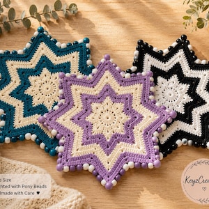 May include: Three handmade star-shaped crochet coasters in teal, purple, and black. Each coaster features a central floral design, a contrasting border, and is edged with decorative beads. The coasters are displayed on a wooden surface.