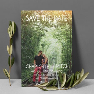 K&ouml;nnte beinhalten: Save-the-Date-Karte mit einem Foto eines Paares, das auf einem Bahngleis in einem Wald steht. Der Text auf der Karte lautet "Save the Date, Charlotte und Mitch heiraten, Sonntag, 22. Oktober 2023, Los Angeles, CA 123456".