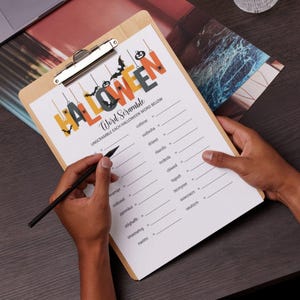 May include: A wooden clipboard holds a Halloween-themed word scramble game. The top of the paper features the word "HALLOWEEN" in orange, black, and green letters with Halloween-themed illustrations. A person is holding a black pen, ready to solve the puzzle.