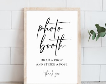 May include: A white sign with a black frame, reading "photo booth" in a cursive font. Below the text, it says "Grab a prop and strike a pose" and "Thank you".