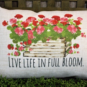 May include: A white pillow with a floral design of pink and red geraniums in a wooden planter. The pillow has the text "LIVE LIFE IN FULL BLOOM." printed on it.