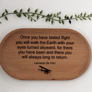 K&ouml;nnte beinhalten: Ein ovales Holztablett mit dem Zitat &bdquo;Sobald du die Flucht gekostet hast...&ldquo; von Leonardo Da Vinci und einer Flugzeugillustration. Eine gr&uuml;ne Ranke befindet sich dar&uuml;ber.