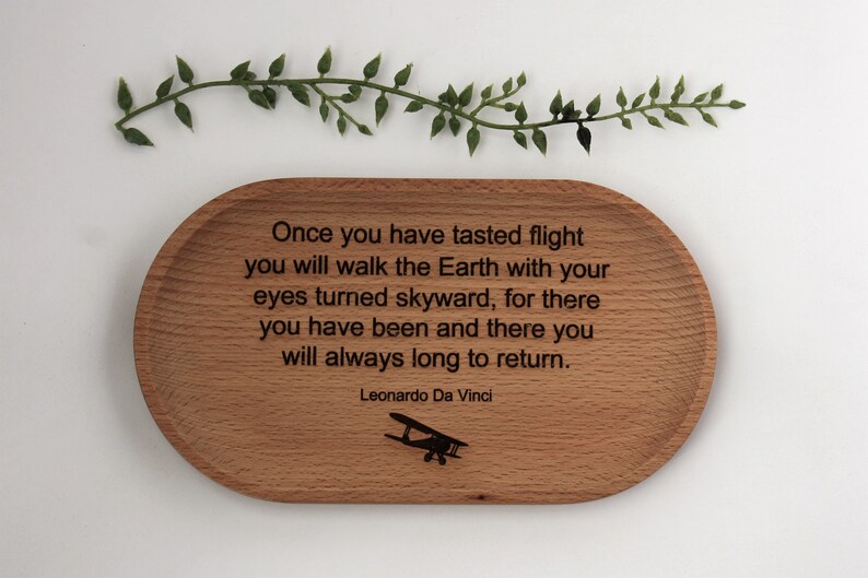 K&ouml;nnte beinhalten: Ein ovales Holztablett mit dem Zitat &bdquo;Sobald du die Flucht gekostet hast...&ldquo; von Leonardo Da Vinci und einer Flugzeugillustration. Eine gr&uuml;ne Ranke befindet sich dar&uuml;ber.
