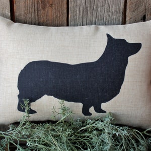 May include: A beige pillow with a black silhouette of a corgi dog printed on it.
