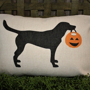 May include: A white cushion with a black silhouette of a dog carrying a jack-o'-lantern. The cushion is on a green surface.