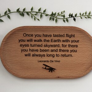 K&ouml;nnte beinhalten: Ein ovales Holztablett mit eingraviertem Text und einer Flugzeugsilhouette. Der Text lautet: &bdquo;Wenn du einmal die Flucht gekostet hast...&ldquo; von Leonardo Da Vinci. Eine gr&uuml;ne, blattreiche Rebe befindet sich &uuml;ber dem Tablett.