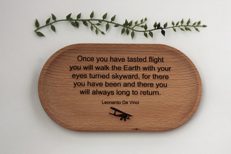 K&ouml;nnte beinhalten: Ein ovales Holztablett mit eingraviertem Text und einer Flugzeugsilhouette. Der Text lautet: &bdquo;Wenn du einmal die Flucht gekostet hast...&ldquo; von Leonardo Da Vinci. Eine gr&uuml;ne, blattreiche Rebe befindet sich &uuml;ber dem Tablett.