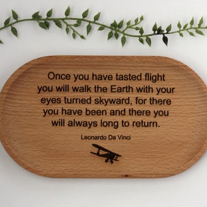 K&ouml;nnte beinhalten: Ovale Holzschale mit eingraviertem Zitat von Leonardo Da Vinci: "Sobald du den Flug gekostet hast..." Enth&auml;lt eine kleine Flugzeuggrafik. Dekorative gr&uuml;ne Zweige oben.
