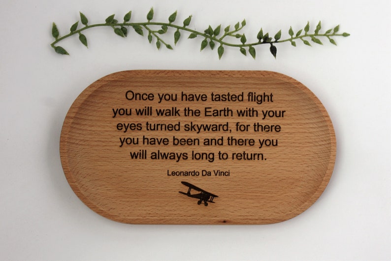 K&ouml;nnte beinhalten: Ovale Holzschale mit eingraviertem Zitat von Leonardo Da Vinci: "Sobald du den Flug gekostet hast..." Enth&auml;lt eine kleine Flugzeuggrafik. Dekorative gr&uuml;ne Zweige oben.