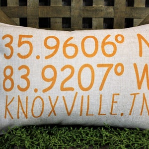 May include: Rectangular linen pillow with orange text: 35.9606° N, 83.9207° W, KNOXVILLE, TN. The pillow has a neutral color and a textured appearance. The text is in a sans-serif font. The pillow is a decorative item.