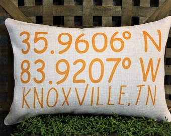 Może przedstawiać: Prostokątna lniana poduszka z pomarańczowym tekstem: 35.9606° N, 83.9207° W, KNOXVILLE, TN. Poduszka ma neutralny kolor i teksturowany wygląd. Tekst jest w czcionce bezszeryfowej. Jest to przedmiot dekoracyjny.