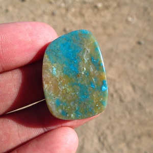 May include: A smooth, oval-shaped cabochon gemstone with a mix of blue, green, and brown colours. The stone has a mottled pattern and a polished finish.