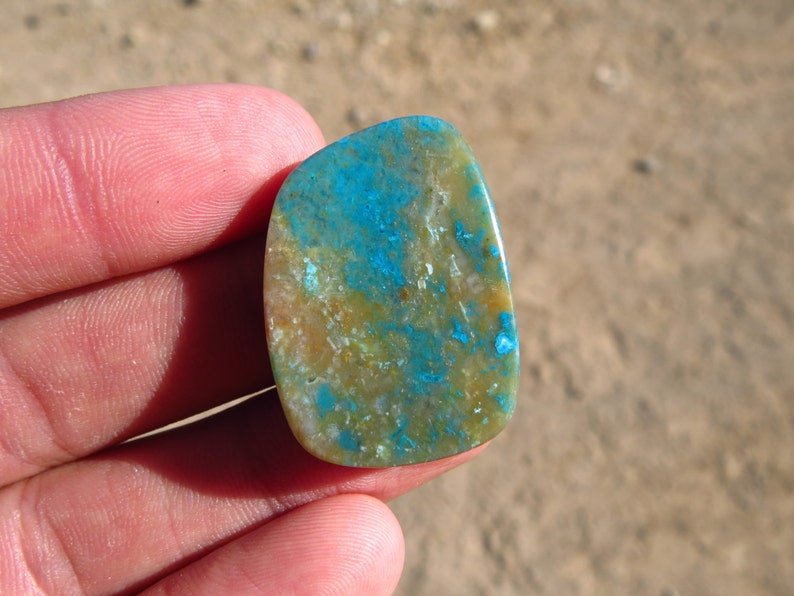 May include: A smooth, oval-shaped cabochon gemstone with a mix of blue, green, and brown colours. The stone has a mottled pattern and a polished finish.