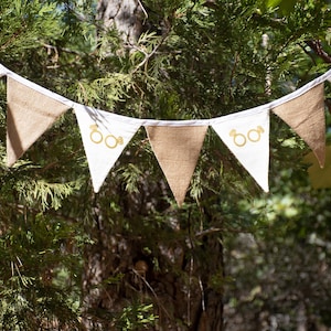 May include: A decorative banner with alternating white and burlap pennants. Each white pennant features a gold ring design. The banner is strung against a backdrop of green foliage and a tree trunk, suitable for a wedding or engagement.