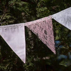 May include: A string of decorative pennant flags in shades of pink and white. Some flags are solid, while others feature a delicate lace design. The flags are strung together and hang against a backdrop of green foliage.