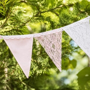 May include: A decorative bunting banner featuring alternating triangular flags in soft pink and white. The flags are crafted from a combination of solid fabric and intricate lace panels, all suspended from a matching fabric trim header.