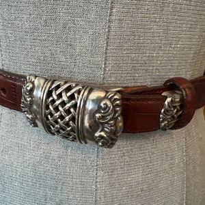 May include: Brown leather belt with a silver buckle. The buckle has a woven centre design with decorative scrollwork on the sides. The belt is displayed against a grey background.