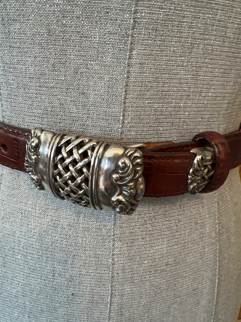 May include: Brown leather belt with a silver buckle. The buckle has a woven centre design with decorative scrollwork on the sides. The belt is displayed against a grey background.