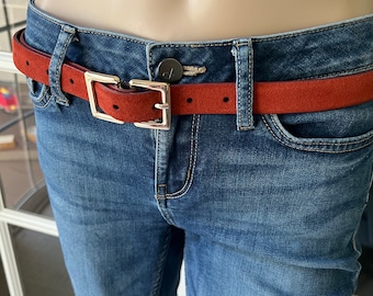 May include: A rust-colored suede belt with a silver-toned square buckle is worn with blue denim jeans. The belt is a thin style, and the buckle is a simple, modern design. The jeans have a classic five-pocket design.