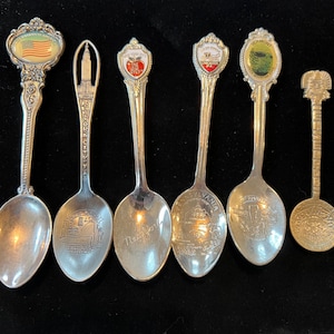 May include: A collection of vintage souvenir spoons. The spoons feature various designs, including the American flag, the Empire State Building, and state emblems. The spoons are made of silver-toned metal and are arranged on a dark surface.