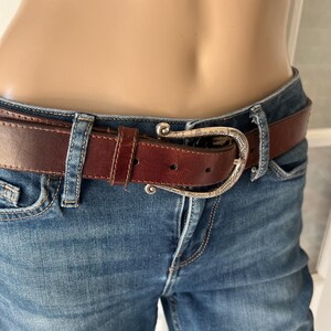 Peut inclure: Une ceinture en cuir marron avec une boucle argent&eacute;e est port&eacute;e avec un jean bleu. La ceinture a une boucle d&eacute;corative et est fix&eacute;e aux passants du jean. Le jean est en denim d&eacute;lav&eacute; moyen.