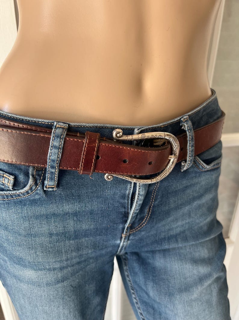 Peut inclure: Une ceinture en cuir marron avec une boucle argent&eacute;e est port&eacute;e avec un jean bleu. La ceinture a une boucle d&eacute;corative et est fix&eacute;e aux passants du jean. Le jean est en denim d&eacute;lav&eacute; moyen.