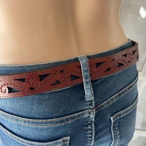 Peut inclure: Une ceinture en cuir marron avec un motif d&eacute;coup&eacute; d&eacute;coratif est port&eacute;e avec un jean bleu. La ceinture pr&eacute;sente une s&eacute;rie de formes g&eacute;om&eacute;triques et courbes d&eacute;coup&eacute;es dans le cuir. Le jean a une conception classique &agrave; cinq poches.