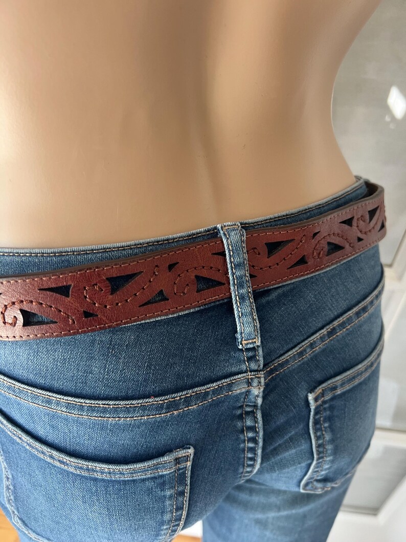 Peut inclure: Une ceinture en cuir marron avec un motif d&eacute;coup&eacute; d&eacute;coratif est port&eacute;e avec un jean bleu. La ceinture pr&eacute;sente une s&eacute;rie de formes g&eacute;om&eacute;triques et courbes d&eacute;coup&eacute;es dans le cuir. Le jean a une conception classique &agrave; cinq poches.