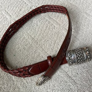 May include: A brown leather belt with a braided design and a silver-coloured buckle. The belt has a textured appearance and a classic style. The buckle features an intricate woven pattern. The belt is approximately 3.2 cm wide.