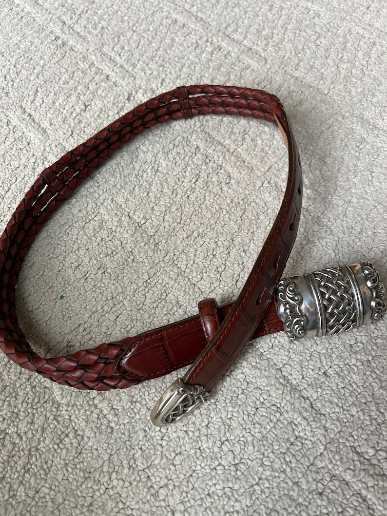 May include: A brown leather belt with a braided design and a silver-coloured buckle. The belt has a textured appearance and a classic style. The buckle features an intricate woven pattern. The belt is approximately 3.2 cm wide.