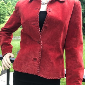 May include: A red suede jacket with a collared neckline and three buttons. The edges are accented with a decorative stitch. The jacket is displayed on a mannequin, with a black top underneath.