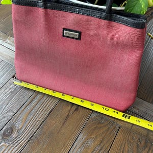 May include: A red tote bag with black handles and trim. The bag has a rectangular shape and a small rectangular tag that reads "JIM THOMPSON". The bag is approximately 13 inches wide.
