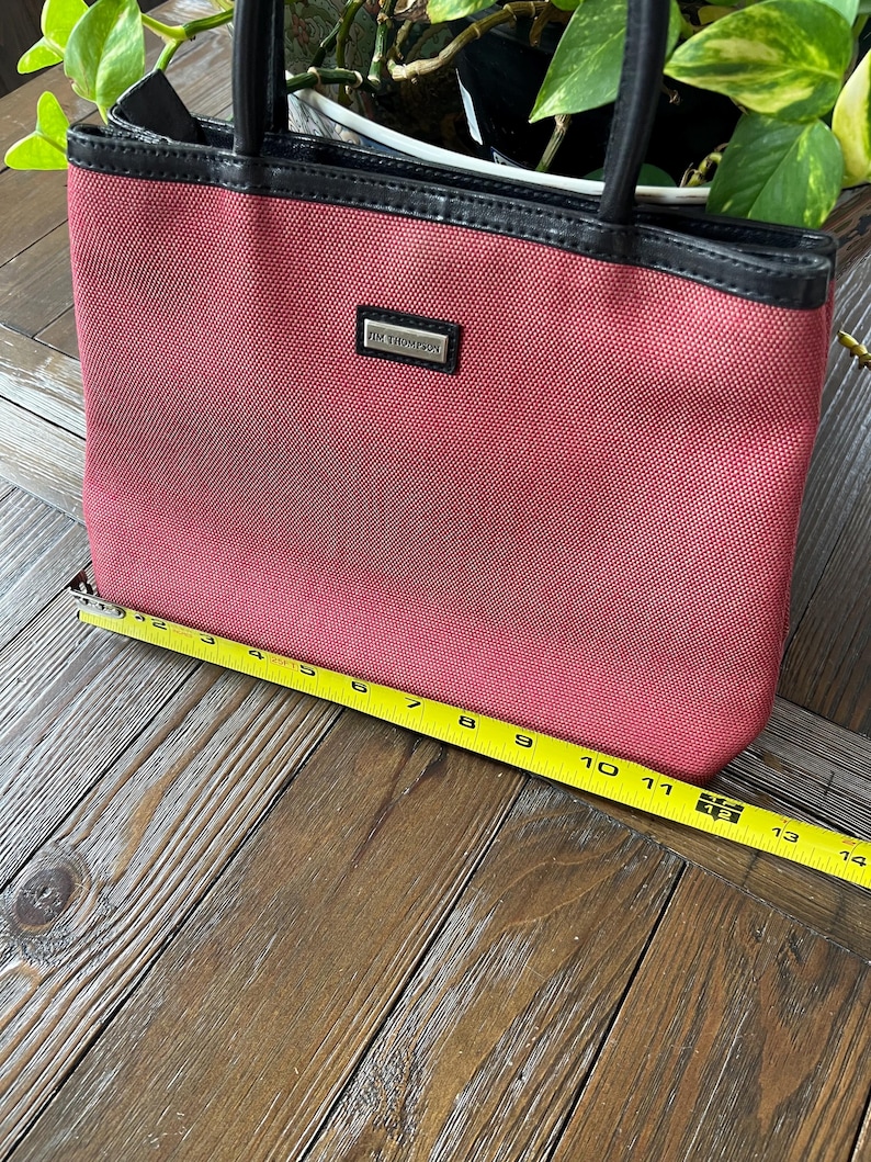 May include: A red tote bag with black handles and trim. The bag has a rectangular shape and a small rectangular tag that reads "JIM THOMPSON". The bag is approximately 13 inches wide.