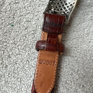 May include: Close-up of a brown leather belt with a silver buckle. The belt has a braided design and a small leather loop. The number "90607" and the letter "S" are stamped on the leather. The buckle is rectangular with a decorative pattern.