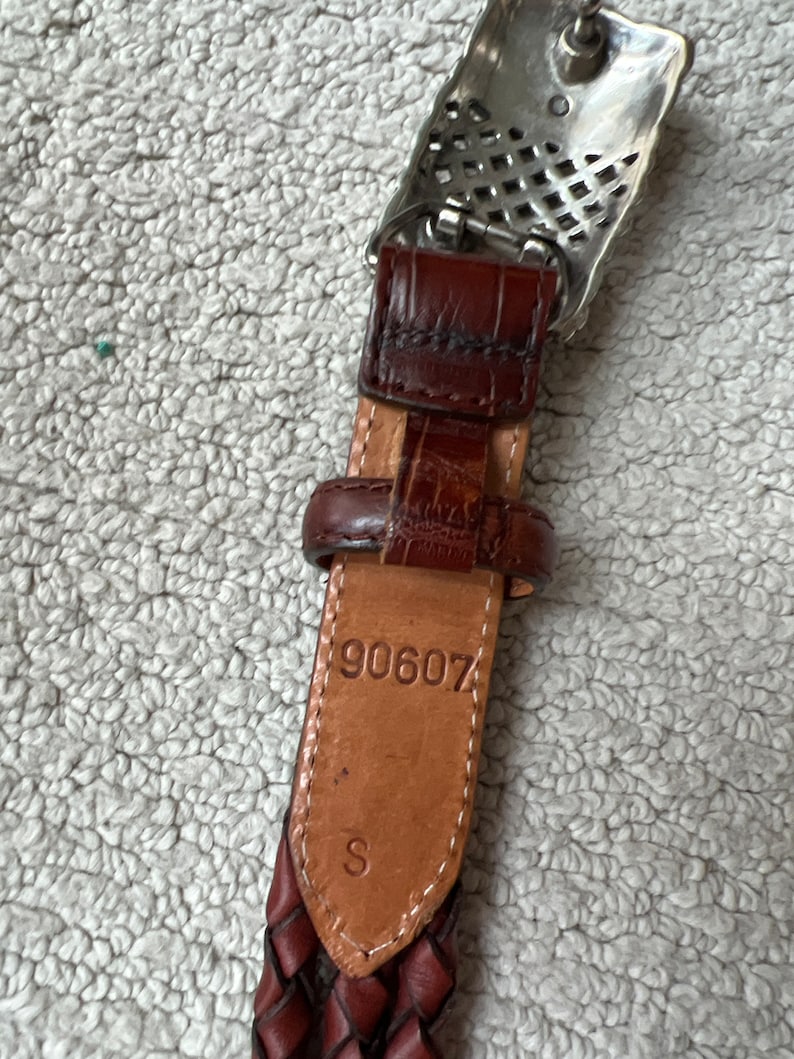 May include: Close-up of a brown leather belt with a silver buckle. The belt has a braided design and a small leather loop. The number "90607" and the letter "S" are stamped on the leather. The buckle is rectangular with a decorative pattern.