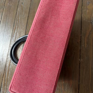 May include: A rectangular, red handbag with a textured fabric. The bag has a black handle and a structured design. The bag is made of a durable material and is suitable for everyday use.