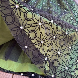 May include: Close-up of a green dress with a black floral lace overlay. The lace is adorned with sparkling embellishments. The dress has a layered design with a tulle underskirt, creating a textured and elegant look. The dress is suitable for special occasions.