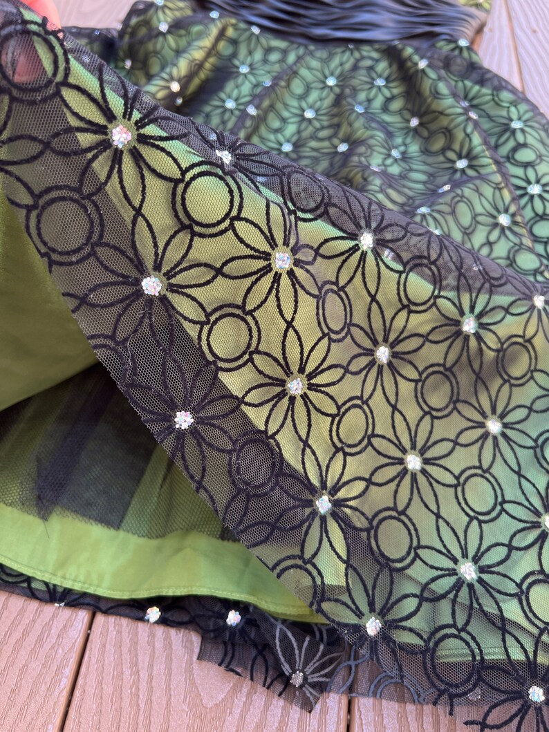 May include: Close-up of a green dress with a black floral lace overlay. The lace is adorned with sparkling embellishments. The dress has a layered design with a tulle underskirt, creating a textured and elegant look. The dress is suitable for special occasions.