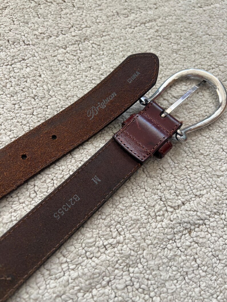 Peut inclure: Une ceinture en cuir marron avec une boucle argent&eacute;e. La ceinture porte la marque "Brighton" et la taille "M". Fabriqu&eacute; en Chine.