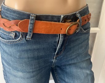May include: A tan leather belt with a braided section and a brass buckle is worn with blue denim jeans. The belt is a warm shade of orange-brown and appears to be made of genuine leather. The jeans are a classic blue wash.
