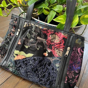 Puede incluir: Un bolso tote patchwork negro y multicolor con dos cremalleras y asas negras. El bolso presenta estampados florales y abstractos en tonos rojos, rosas y azules. El bolso está sobre una superficie de madera.
