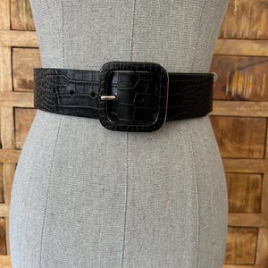 Può includere: Una cintura nera con fibbia quadrata, esposta su un manichino. La cintura presenta un motivo strutturato a forma di coccodrillo. La fibbia è anch'essa nera con un perno in metallo color argento.