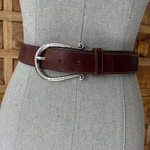 Peut inclure: Une ceinture en cuir marron avec une boucle d&eacute;corative de couleur argent&eacute;e. La boucle pr&eacute;sente un design orn&eacute; avec des bords incurv&eacute;s. La ceinture est pr&eacute;sent&eacute;e sur un mannequin. C'est un accessoire de mode.