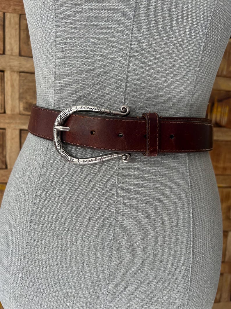 Peut inclure: Une ceinture en cuir marron avec une boucle d&eacute;corative de couleur argent&eacute;e. La boucle pr&eacute;sente un design orn&eacute; avec des bords incurv&eacute;s. La ceinture est pr&eacute;sent&eacute;e sur un mannequin. C'est un accessoire de mode.