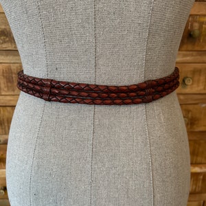 May include: A brown leather belt with a braided design. The belt is wrapped around a mannequin. The belt has three braided strands and a dark brown colour. The belt is a fashion accessory.