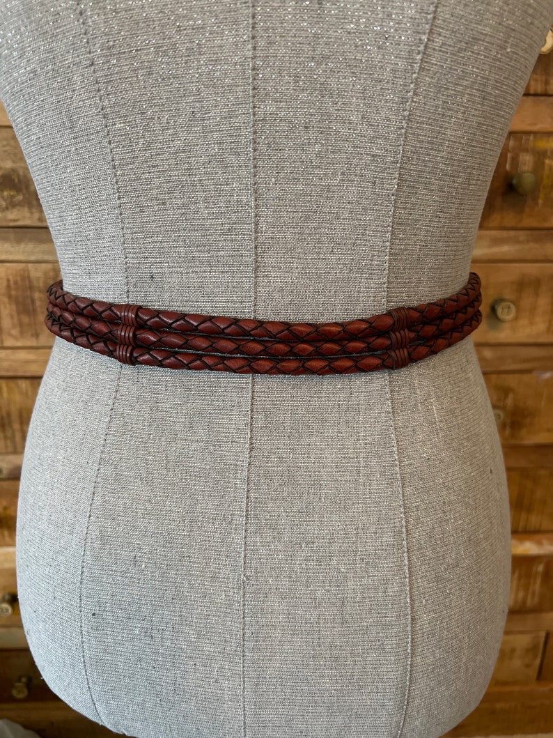 May include: A brown leather belt with a braided design. The belt is wrapped around a mannequin. The belt has three braided strands and a dark brown colour. The belt is a fashion accessory.